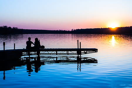 silhouette photo of man and woman sitting on dock near blue body of water during golden hour thumbnail