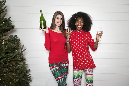 two women wearing red shirts holding wine bottle and two champagne flutes standing near wall thumbnail
