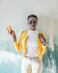 woman wearing yellow zip-up jacket, white crew-neck top, and gray pants standing in front of teal painted wall while holding brown ice cream cone thumbnail