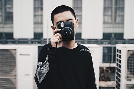 man standing behind the building while holding black Fujifilm Instax camera thumbnail