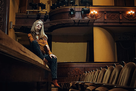 woman sitting on brown wooden theater stage beside chairs thumbnail
