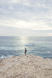 man wearing grey shirt standing on cliff facing sea water thumbnail