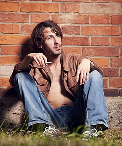man wearing brown leather jacket and blue jeans sitting on grass near brick wall thumbnail