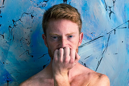 man leaning on blue concrete wall biting his left hand thumbnail
