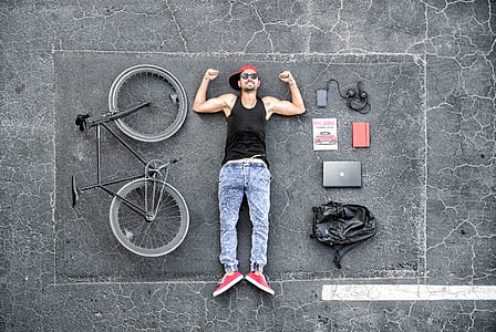 man wearing black tank top and blue denim jeans lying on road beside bike and laptop thumbnail
