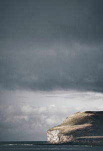 cliff beside body of water under cloudy sky thumbnail