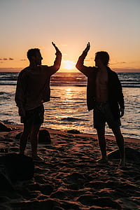 silhouette of two men doing high five standing on sandy beach during sunset thumbnail