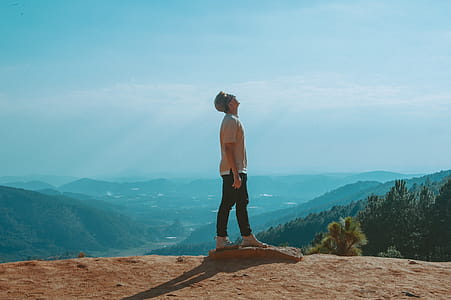 Man Wearing Beige Shirt and Black Pants Looking Up With Blue Sky and Mountains in the Background thumbnail