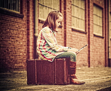 woman wearing green, red, and brown long-sleeved shirt, blue denim jeans and brown leather cowboy boots sitting on brown luggage bag thumbnail
