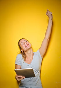 Young Happy Woman Holding An iPad With One Hand Up Against Yellow Wall thumbnail