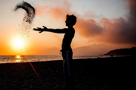 silhouette photography of man throwing sand near body of water during golden hour thumbnail