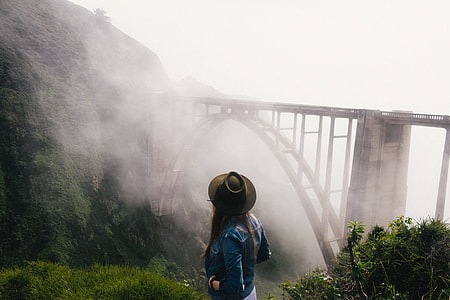 woman in cowboy hat and blue denim jacket standing on cliff with a view of concrete train bridge covered with fogs thumbnail