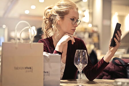 A young blond woman wearing a satin dress looks at her mobile phone while enjoying a glass of wine thumbnail