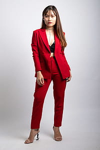 Woman Wearing Red Coat and Dress Pants Standing in Front of White Wallpaper thumbnail