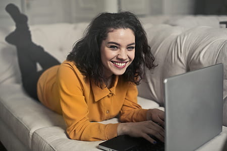 A young woman in yellow button-up shirt and black leggings smiling while working on her laptop on the white couch thumbnail