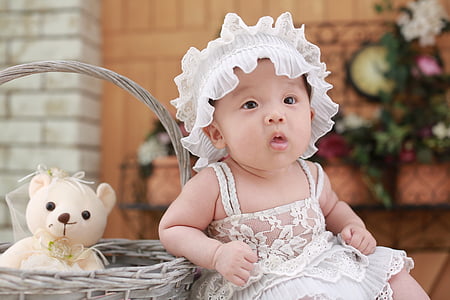 baby wearing white dress beside basket with teddy bear sitting on table outdoors thumbnail