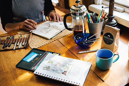 person performing art on notebook on table beside coffee press and coloring kit thumbnail