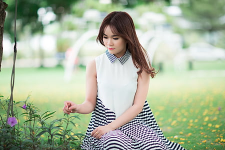 black haired woman wearing white and black striped sleeveless dress sitting on green grass field thumbnail