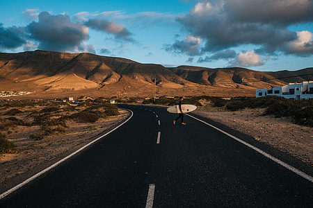 person carrying surfboard crossing road alone with mountain background during daytime thumbnail