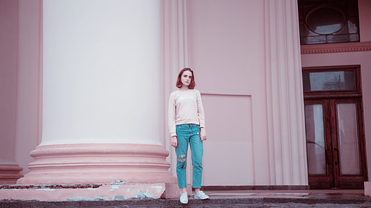 woman wearing white sweater , teal jeans and white sneakers standing beside building pillar thumbnail