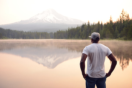 man in white shirt and blue jeans standing near lake and body of mountain during daytime thumbnail