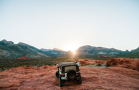 white and black SUV above brown rock formation with man wearing blue shirt standing beside SUV surrounded by black mountains during sunrise thumbnail