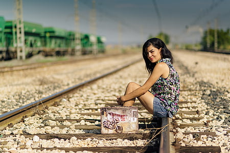 woman wearing black and multicolored sleeveless top and blue denim shorts outfit sitting on train railways under blue sky during daytime thumbnail