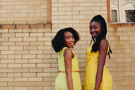 two girls wearing yellow sleeveless dresses smiling near brown brick wall thumbnail