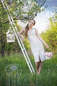 woman wearing white lace dress holding basket leaning on white wooden ladder with birdcage thumbnail