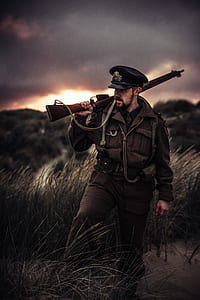 man wearing military uniform holding rifle on his shoulder standing in tall grass field thumbnail
