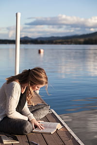 woman sitting on dock while reading book during daytime thumbnail