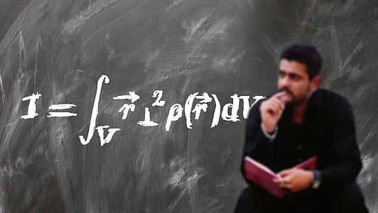man holding opened book while sitting with black chalkboard in background thumbnail