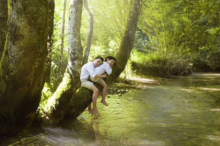 two boys sitting on tree branch above stream during day thumbnail