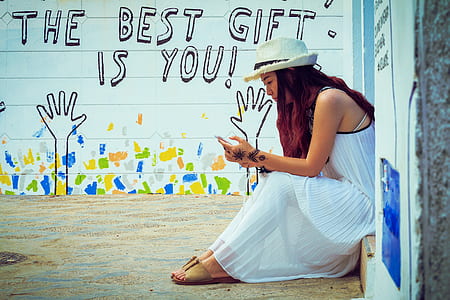 woman in white sleeveless dress sitting on doorway and using mobile phone during daytime thumbnail