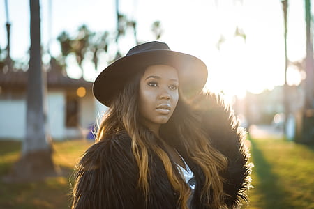 woman wearing black fur coat and black fedora hat standing near trees thumbnail