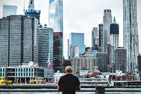 man wearing black shirt leaning on gray metal railing facing city buildings during daytime thumbnail