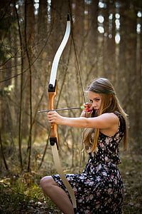 woman in black floral dress holding bow and arrow kneeling in forest thumbnail