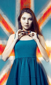 woman in blue scoop-neck sleeveless dress holding bow in front of her neck with Union flag in background thumbnail