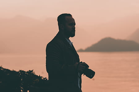 man standing on mountain near body of water holding black DSLR camera thumbnail
