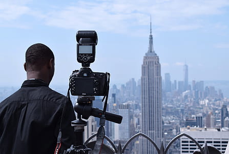 man in black shirt standing beside a camera with tripod capturing the grey high-rise building thumbnail