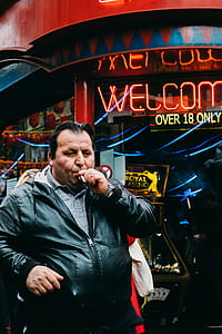 man wearing black leather jacket smoking beside neon welcome sign thumbnail