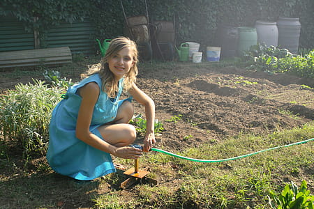 A young blonde woman in shirt dress holding a plant watering fountain in the garden thumbnail