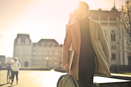 Young bearded male backlit by the sun, wearing black turtleneck top with long overcoat and eyeglasses carrying a laptop,as he walks on the street thumbnail