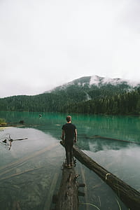 man in black shirt standing on brown tree log on lake during daytime thumbnail