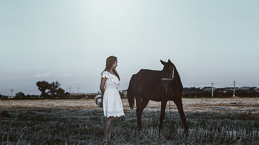 woman in white dress standing near brown horse in open field under gray sky during daytime thumbnail