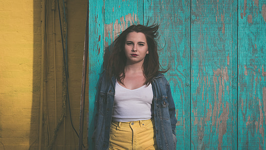 shallow focus photography of woman in white tank top and yellow jeans wearing blue denim jacket leaning on green wooden wall during daytime thumbnail