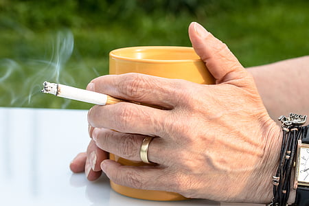 person holding lighted cigarette stick and mug on top of white table thumbnail