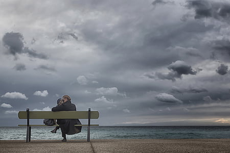 woman sitting on bench facing body of water under cloudy sky thumbnail