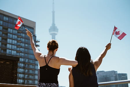 two women holding mini flags of Canada facing CN Tower in Toronto during daytime thumbnail