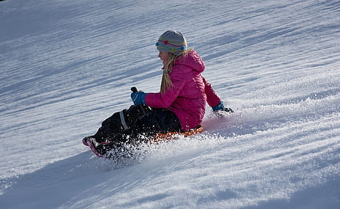 woman wearing pink zip-up hooded bubble jacket sits on orange sled on top of snow thumbnail